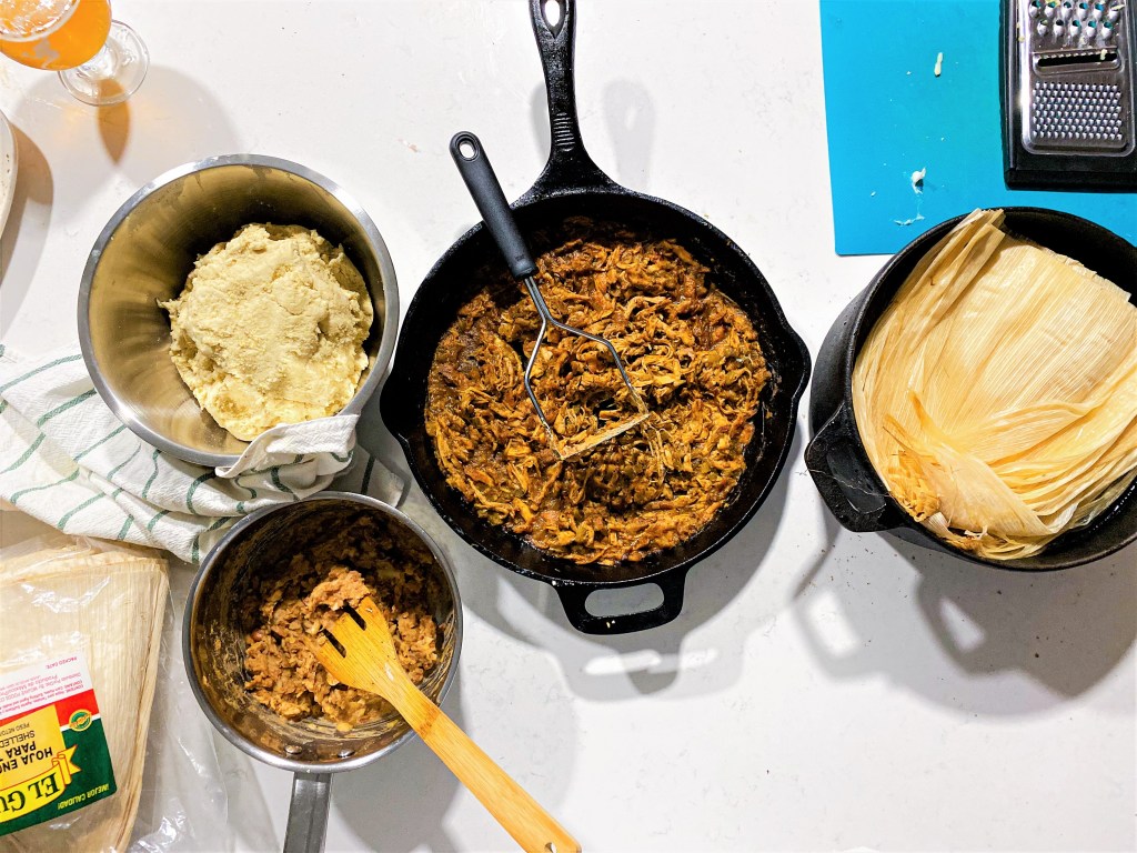 Tamale ingredients like masa, corn husks, pork, and red beans laid out on a table, ready to be assembled.